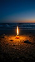 Single candle flame glowing on sandy beach at twilight with ocean waves and dramatic sunset sky creating peaceful meditation atmosphere.