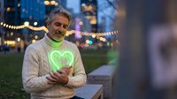 Mature Caucasian man wearing green heart-shaped necklace in urban evening setting with bokeh lights, expressing love and romance during city date.