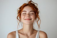 Young woman with red hair and closed eyes expressing joy and serenity against light background, wearing white tank top and delicate necklace, natural makeup and messy updo hairstyle.