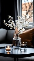White cherry blossom branches in glass vase alongside glowing candles on round coffee table, creating cozy home atmosphere against dark window background.