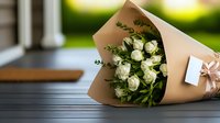 Fresh white roses wrapped in kraft paper with decorative bow and gift card lying on wooden surface against blurred garden background. Romantic floral gift concept.