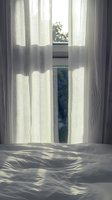 Sunlight streaming through sheer white curtains onto rumpled bedding creates peaceful morning atmosphere with window view of nature and soft shadows.