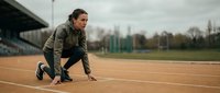 Young Caucasian female athlete in starting position on outdoor running track, focused and ready to sprint, wearing olive sportswear on cloudy day.