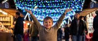 Young boy with arms raised celebrating at festive Christmas market with colorful lights overhead and holiday shopping stalls in background during evening.