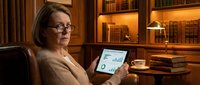 Mature woman with glasses holding tablet displaying financial charts in warm-lit home office with bookshelves, studying investment data or business analytics.