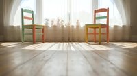 Colorful wooden chairs facing each other in bright sunlit room with sheer curtains, casting long shadows on wooden floor, symbolizing dialogue and connection.