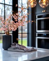 Pink cherry blossoms in modern black ribbed vase on marble countertop, gray linen napkins, contemporary kitchen interior with pendant lights and window view.
