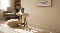 Cozy minimalist living room with wooden stool, knit pouf, and mindfulness wall art in warm natural light, promoting calm and wellness.