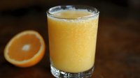 Fresh squeezed orange juice in clear glass with frothy top and half orange slice on dark wooden surface, natural morning beverage photography.