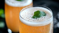 Fresh cold beer with white foam head and mint leaf garnish in glass, bubbles rising through golden amber liquid against dark background.