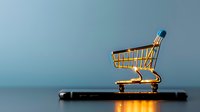 Small golden shopping cart placed on black smartphone against blue background, representing mobile commerce and digital retail solutions.