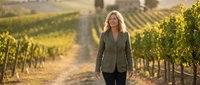 Professional blonde businesswoman walking through vineyard rows during golden hour sunset, wine industry agriculture concept for corporate marketing.