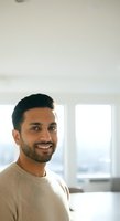 Portrait of a smiling South Asian male professional in a beige sweater, standing indoors near bright windows with cityscape views for corporate and lifestyle use.