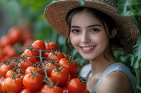 Young Asian woman in straw hat smiling next to fresh ripe tomatoes on vine in greenhouse garden, representing healthy lifestyle and organic farming.