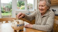 Smiling senior Caucasian woman in beige cardigan putting coins into piggy bank labeled pension at kitchen table, retirement savings concept for financial planning.