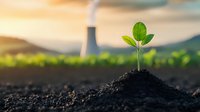 Young green seedling growing from fertile soil with industrial chimney emitting smoke in blurred background, environmental concept showing contrast between nature and pollution.