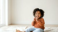 Young child with curly dark hair and medium skin tone wearing rust-colored shirt and jeans, sitting on bed in bright bedroom with white walls and natural window light, smiling and laughing.