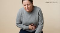 Middle-aged Caucasian woman experiencing severe abdominal pain holding stomach in gray sweater expressing discomfort and digestive health issues.