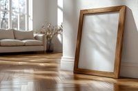 Empty wooden picture frame leaning against white wall in modern living room with herringbone parquet floor, sofa and natural light casting shadows.