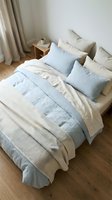 Cozy bedroom interior with neatly arranged bed featuring light blue and cream linen bedding, decorative pillows, wooden nightstand, and soft curtains for home decor and interior design inspiration.