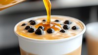 Golden caramel syrup pouring into creamy bubble tea topped with fresh black tapioca pearls, close-up shot showing layered milk tea drink.