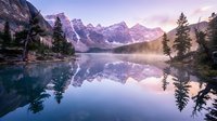 Serene mountain lake with perfect reflections of snow-capped peaks and evergreen trees during golden hour sunrise, creating peaceful wilderness landscape.