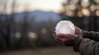 Hands gently holding glowing crystal sphere with warm light emanating from within against blurred mountain landscape at sunset creating magical atmosphere.