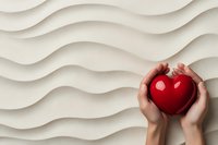 Hands gently holding a shiny red heart against a soft wavy white textured background, symbolizing love, care, and health concepts.