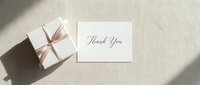 Elegant thank you card with a white gift box tied with a pink ribbon on a neutral linen background, soft natural light and shadows.