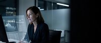 Caucasian female professional in dark business blazer analyzing financial data and stock market charts on computer monitor in modern office environment at night.
