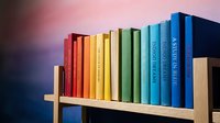 Colorful books arranged on wooden shelf creating rainbow spectrum from red to blue spines against gradient background for education and learning concepts.