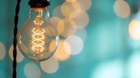 Vintage Edison bulb with spiral filament glowing warmly against soft turquoise background with golden bokeh lights creating cozy atmospheric lighting.
