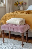 Cozy bedroom corner with pink tufted ottoman, white towel, and mustard yellow bedding. Fresh flowers in vase create welcoming atmosphere for interior design.