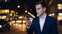 Young professional businessman using smartphone on city street at night with bokeh lights background for mobile technology and urban lifestyle concepts.