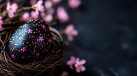 Decorative black Easter egg with colorful star pattern in nest surrounded by delicate pink spring blossoms against dark moody background. Creative holiday composition.