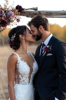 Romantic newlywed couple sharing intimate moment under rustic wooden arch decorated with purple and burgundy flowers during golden hour sunset wedding ceremony.
