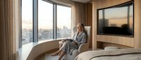 Young professional woman in gray robe relaxing by curved window seat reading tablet in modern luxury hotel bedroom with city skyline view at sunset.