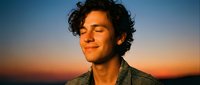 Young Hispanic man with curly hair smiling peacefully with eyes closed during golden hour sunset, expressing joy and serenity in warm evening light.