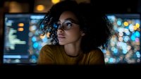 Young African American woman with curly hair and glasses in yellow sweater looking thoughtfully at city lights through window at night.