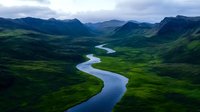 Meandering river flows through verdant mountain valley with dramatic peaks and moody overcast sky, creating natural curves across lush green landscape.