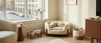 Cozy modern living room interior with comfortable armchair, wooden furniture and large windows overlooking urban street. Warm neutral tones create inviting atmosphere.