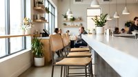 Modern cafe interior with wooden chairs at counter seating, pendant lighting, potted plants, and blurred customers in background, suitable for hospitality and restaurant design projects.