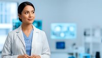 Professional female doctor in white coat standing in modern medical facility with blue medical equipment background for healthcare presentations.