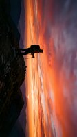 Silhouetted figure standing on rocky cliff edge against dramatic fiery sunset sky with vibrant orange and red clouds, suitable for adventure, inspiration, and motivational content.
