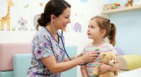 Female pediatric nurse examining young Caucasian girl with stethoscope in colorful children's medical office. Child holds teddy bear during checkup visit.