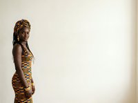 Young woman with dark skin tone and blonde braided hair wearing a gold and black patterned sleeveless dress, standing against neutral background for portrait and fashion use.