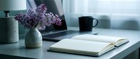 Cozy home office workspace with purple lilac flowers in vase, open notebook, laptop and coffee mug on desk creating peaceful work environment atmosphere.
