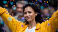 Young mixed race woman with curly hair smiling and celebrating with arms raised wearing yellow jacket while colorful confetti falls around her in joyful celebration moment.