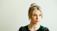 Young woman with blonde hair styled in an updo, fair complexion, wearing a dark green patterned top against a neutral background. Suitable for portrait, lifestyle, and professional use.