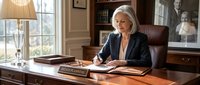 Senior female executive with gray hair and light complexion, wearing dark blazer, writing at wooden desk in upscale office with leather chair, lamp, books, and bookshelf. Corporate professional environment.